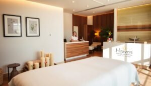 Soft natural lighting bathes an inviting spa interior, revealing a serene scene of relaxation. In the foreground, a cozy massage table with pristine white linens, surrounded by artfully arranged candles and fresh flowers. On the walls, framed nature-inspired artwork and wood-paneled accents create a calming ambiance. The middle ground showcases a sleek reception desk adorned with the "Heaven Massage and Wellness" branding, where a friendly attendant greets arriving clients. Beyond, a tranquil lounge area with plush seating and a soothing water feature provides a perfect space for pre- or post-treatment relaxation. Altogether, an atmosphere of luxurious indulgence and rejuvenation.